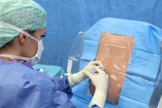 Szenenbild: eine Ärztin in OP-Kleidung injiziert etwas in das Operationsfeld, das von der sterilen Abdeckung des restlichen Patientenkörpers umgeben ist Szenenbild: eine Ärztin in OP-Kleidung injiziert etwas in das Operationsfeld, das von der sterilen Abdeckung des restlichen Patientenkörpers umgeben ist