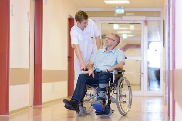 ein Pfleger spricht mit einem Patienten, der eine Fuß-Orthese trägt und in einem Rollstuhl sitzt