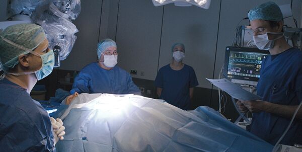 Szenenfoto aus dem Operationssaal, vier Mitglieder des Operationsteams um den zu operierenden Patienten