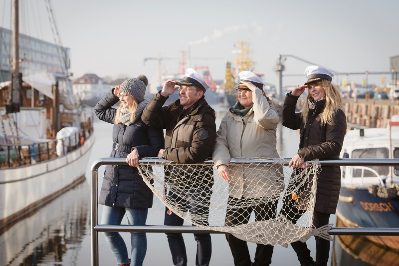 Vier Mitarbeitende der Pflegeakademie mit Kapitänsmützen, die in einem Kutterhafen an einem Geländerstehen, über dem ein Fischernetz hängt