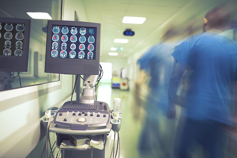 Symbolbild: Überwachungsmonitor in einem Krankenhausflur, daneben verschwommen in Bewegung ein blau gekleideter Pfleger