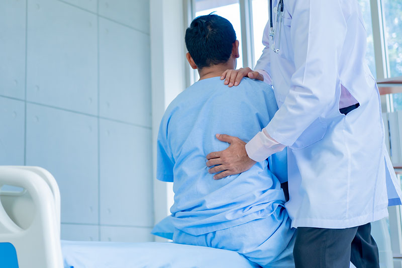 Schmuckbild: ein Arzt untersucht den schmerzenden Rücken eines Patienten, auf einem Krankenhausbett sitzt