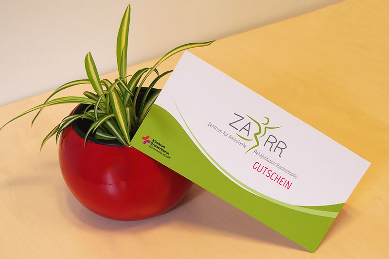 Geschenkgutschein des ZARR Zentrum für Ambulante Rehabilitation Reinkenheide, der an einer kleinen Grünlilie in einem roten Blumentopf lehnt