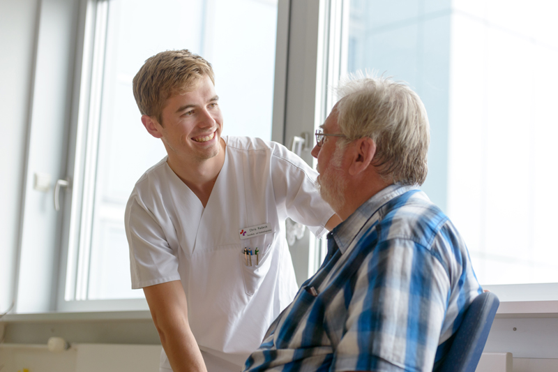 Ein Pfleger spricht mit einem älteren Patienten; verlinkt zur 'Klinik für Akutgeriatrie und geriatrische Frührehabilitation'
