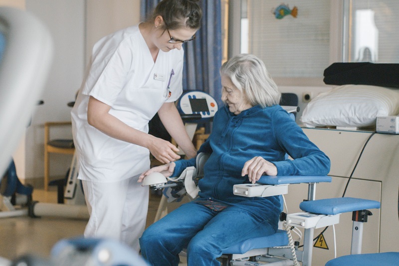 Szenenfoto aus der Klinik für Akutgeriatrie, eine Pflegerin hilft einer sitzenden Patientin