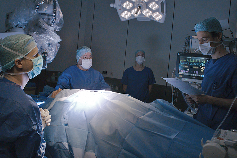 Szenenfoto aus dem Operationssaal, vier Mitglieder des Operationsteams um den zu operierenden Patienten