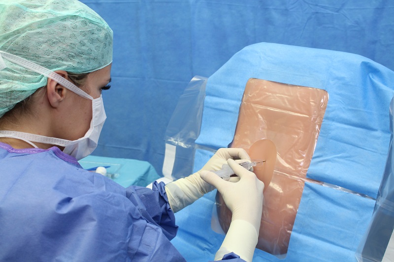 Szenenbild: eine Ärztin in OP-Kleidung injiziert etwas in das Operationsfeld, das von der sterilen Abdeckung des restlichen Patientenkörpers umgeben ist