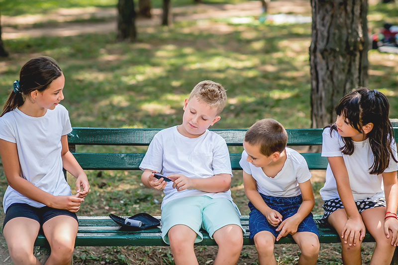 Symbolbild: Ein Junge auf einer Parkbank testet seinen Blutzuckerspiegel, drei andere Kinder schauen freundlich und interessiert zu