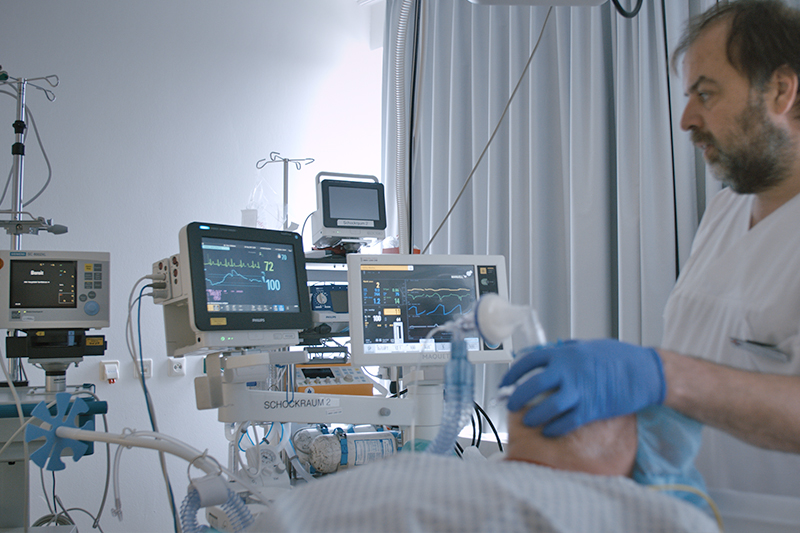 Ein Arzt am Bett eines beatmeten Patienten, im Hintergrund Überwachungsmonitore