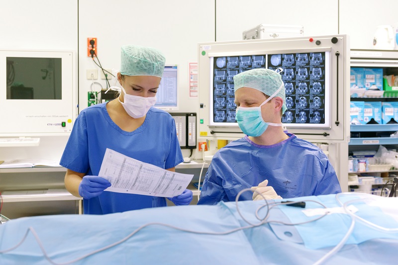 Szenenfoto: ein Mann und eine Frau in OP-Kleidung und mit Mund-Nasen-Schutz, Handschuhen und Kopfhaube blicken im Operationssaal gemeinsam auf einen Bogen aus einer Krankenakte
