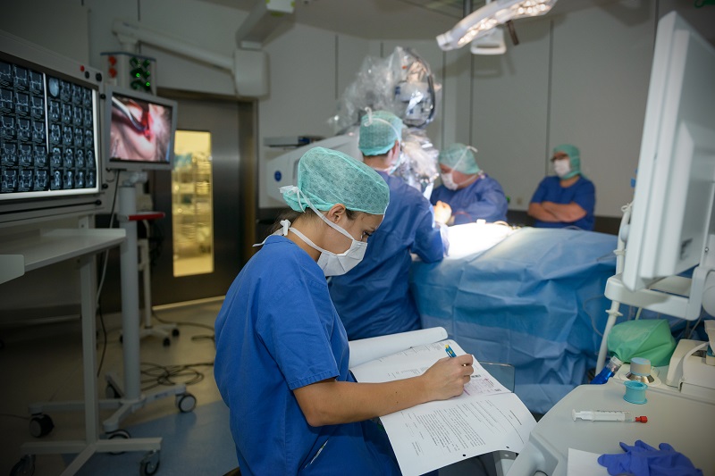 Szenenfoto aus dem Operationssaal, Operateure und Monitore im Hintergrund, im Vordergrund eine Mitarbeiterin, die das Protokoll der Operation schreibt