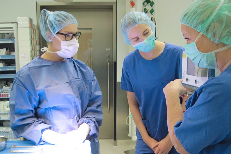 Szenenbild aus der OTA-Ausbildung, eine Mitarbeiterin erklärt zwei Auszubildenden ein Gerät, alle tragen blaue OP-Kleidung, Mund-Nasen-Schutz und Hauben