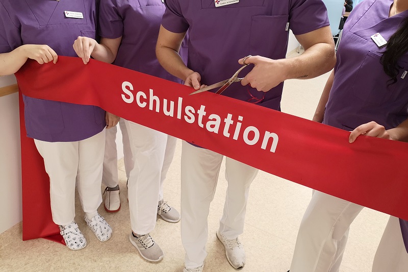 Eröffnung der Schulstation: Pflege-Auszubildende halten und zerschneiden ein breites rotes Band