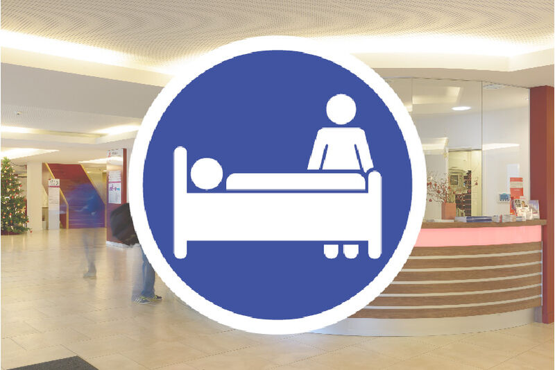Foto der Eingangshalle des Klinikums; davor ein blau-weißes Schild mit Piktogramm eines Patienten im Bett und einer Besucherin