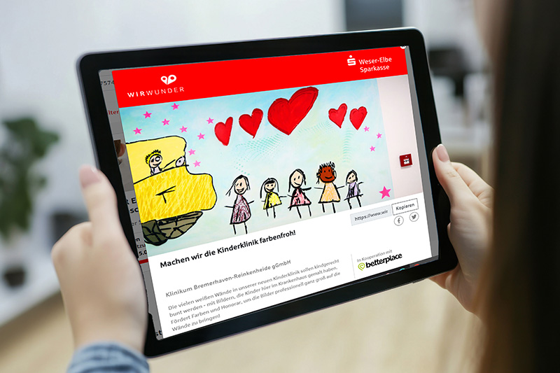 Zwei Hände halten ein Tablet, auf dem die Startseite des Spendenprojekts für die Kinderklinik auf WirWunder zu sehen ist