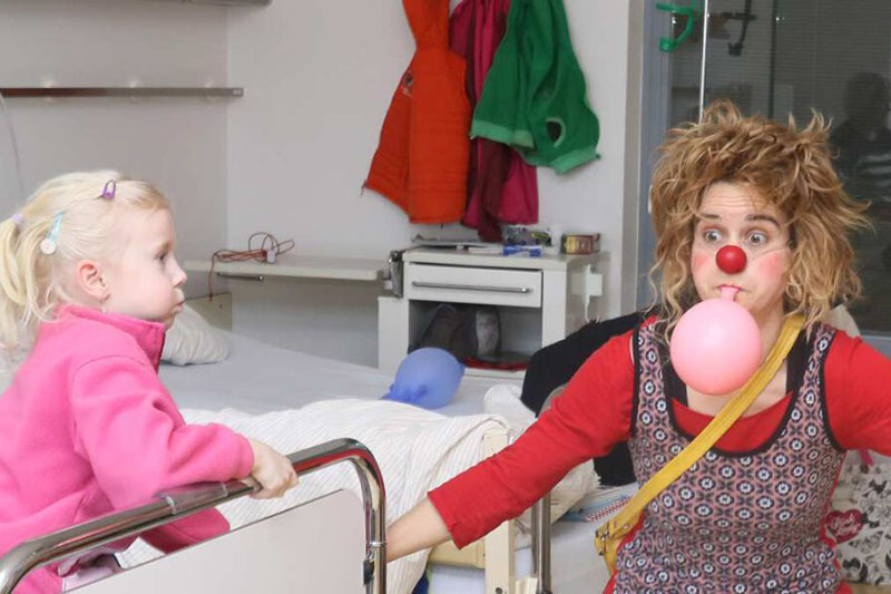 Szenenfoto von einer Klinikclownin mit rosafarbenem Luftballon und einem Kind im Krankenhausbett (verwendet mit freundlicher Genehmigung durch Bremer Klinikclowns e.V.)