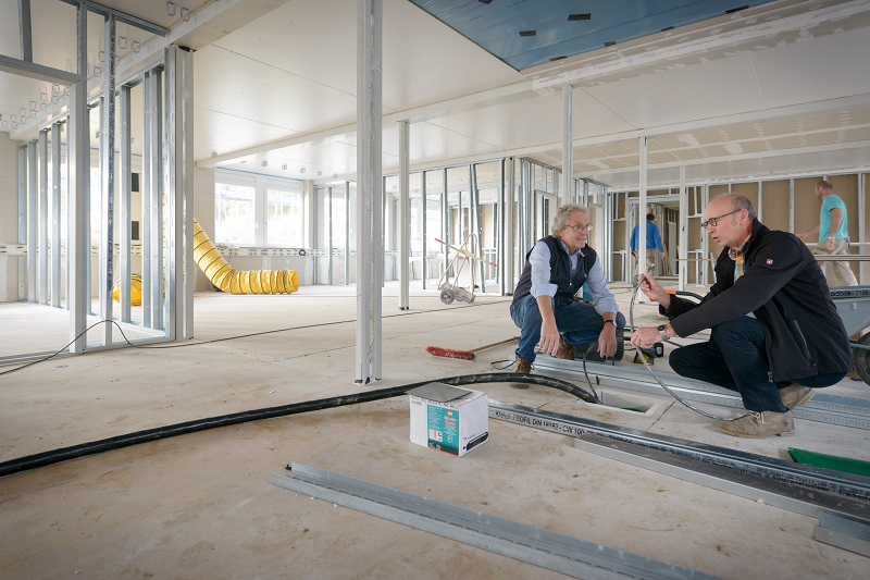 Zwei Mitarbeiter des Architekten- und Planungsteams in einem der Module für die neue Kinderklinik hocken auf dem Boden und betrachten zu verlegende Kabel